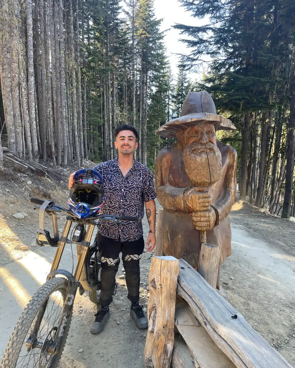 Jaime with his mountain bike beside a carved wooden gnome on a forest trail